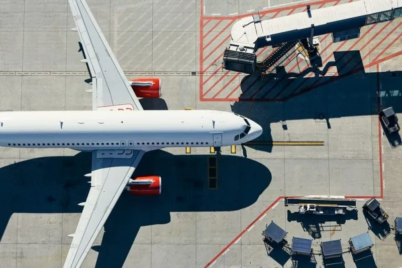 Overhead view of a commercial airplane parked at an airport gate, with a jet bridge connected to the front door, ground service vehicles nearby, and the plane casting a large shadow on the tarmac.