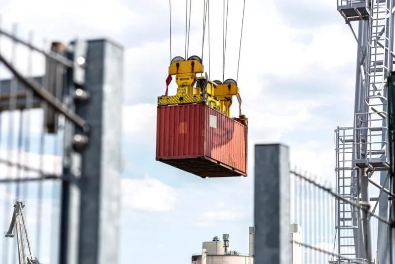 Red shipping container suspended in the air by a crane, being lifted in an industrial port area with metal structures and fencing in the foreground.