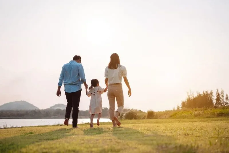 Two adults and a child holding hands while walking barefoot across grass near a body of water at sunset.
