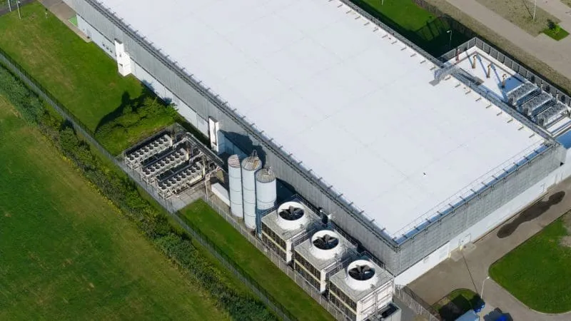 Aerial view of a large industrial warehouse with rooftop equipment, including ventilation units and cooling systems, surrounded by grassy areas.