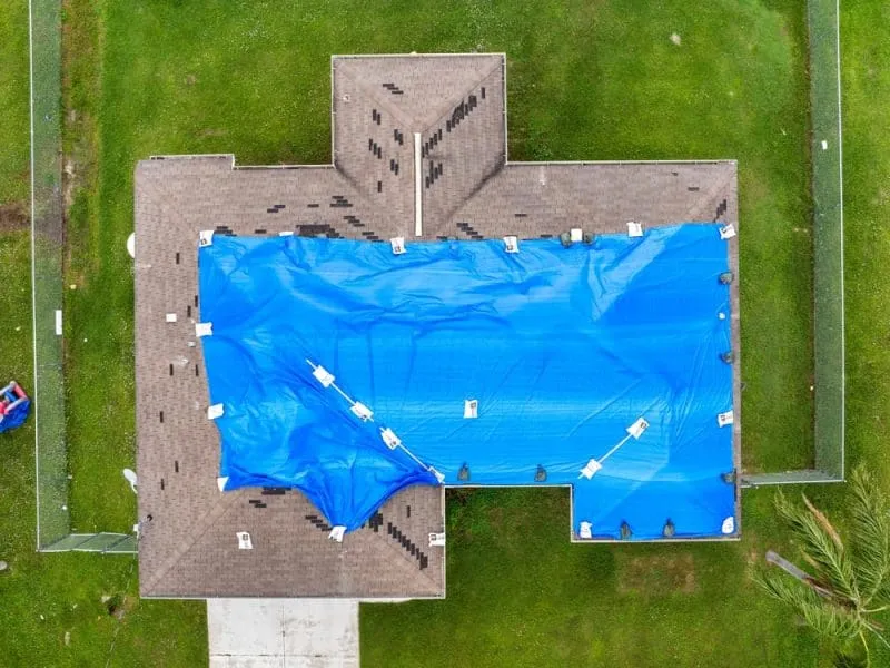 Aerial view of a house roof covered with a blue tarp secured by weights, likely for storm or leak protection, surrounded by a grassy yard.